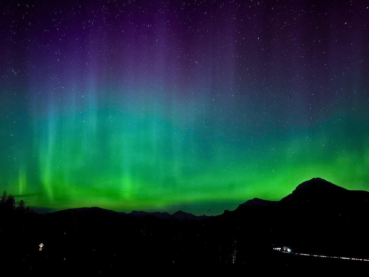 Northern lights with vibrant green and purple hues dancing across a starry night sky above a dark mountain silhouette.