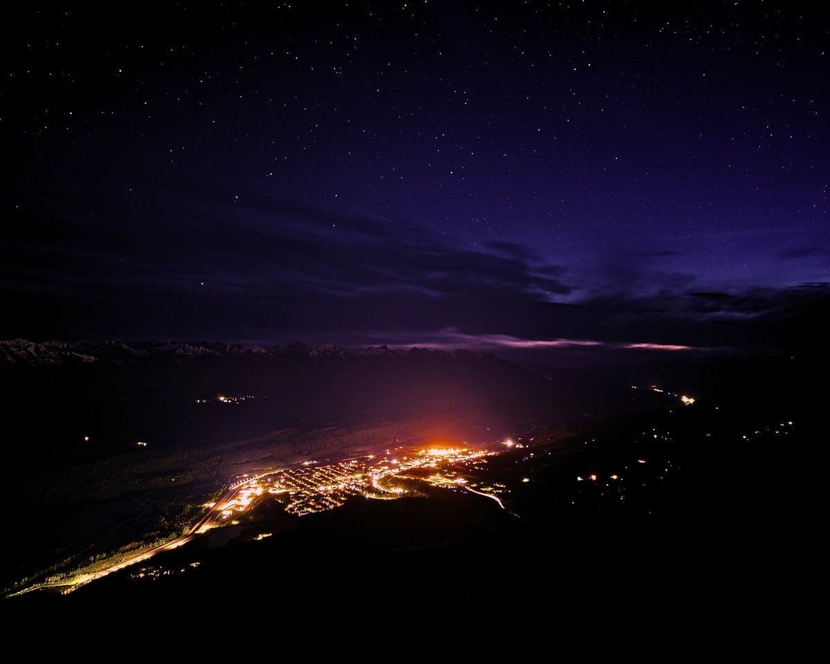 Night landscape photograph showing a starry purple and blue sky over a dark mountainous region with a glowing town below.