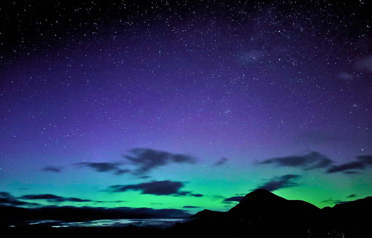 A night sky scene with a star-filled purple and blue sky, green aurora lights near the horizon, and dark mountain silhouettes along the bottom.